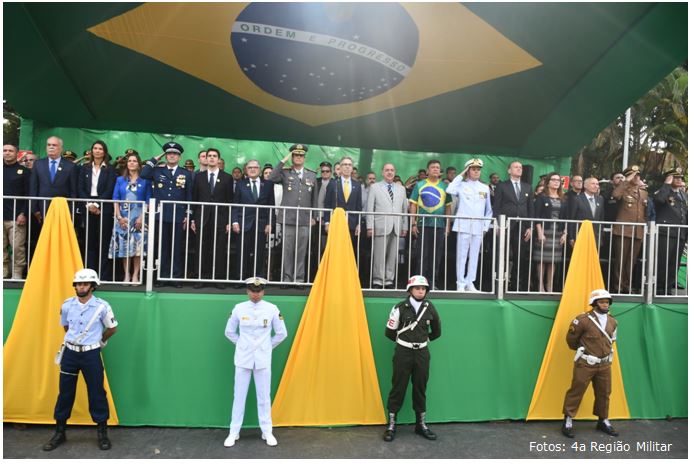 Foto retangular, horizontal, colorida de pessoas reunidas em cima de um palanque decorado das cores verde e amarelo e uma bandeira do Brasil ao topo. Além de quatro militares uniformizados em pé no chão.