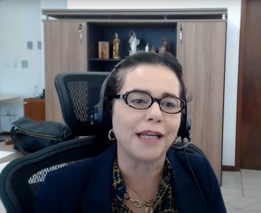 Fotografia horizontal mulher de óculos sentada em gabinete, com fone de ouvido, participando de evento virtual.