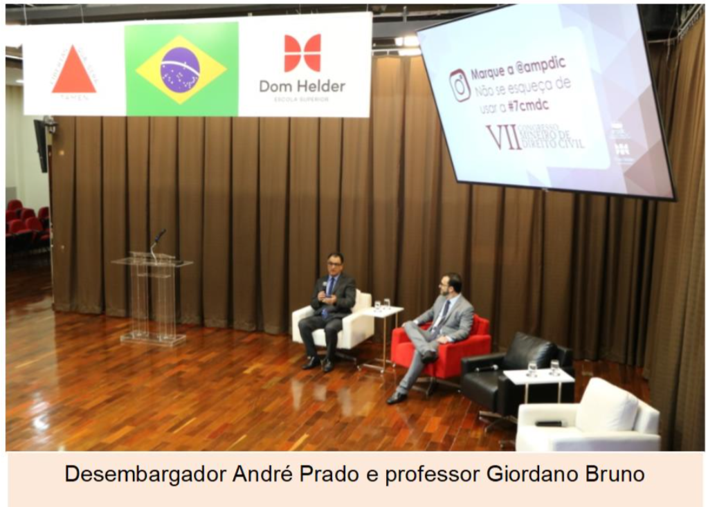 Fotografia horizontal colorida dois homens sentados no palco de um evento, um dos homens falando ao microfone. Legenda: Desembargador André Prado e professor Giordano Bruno.