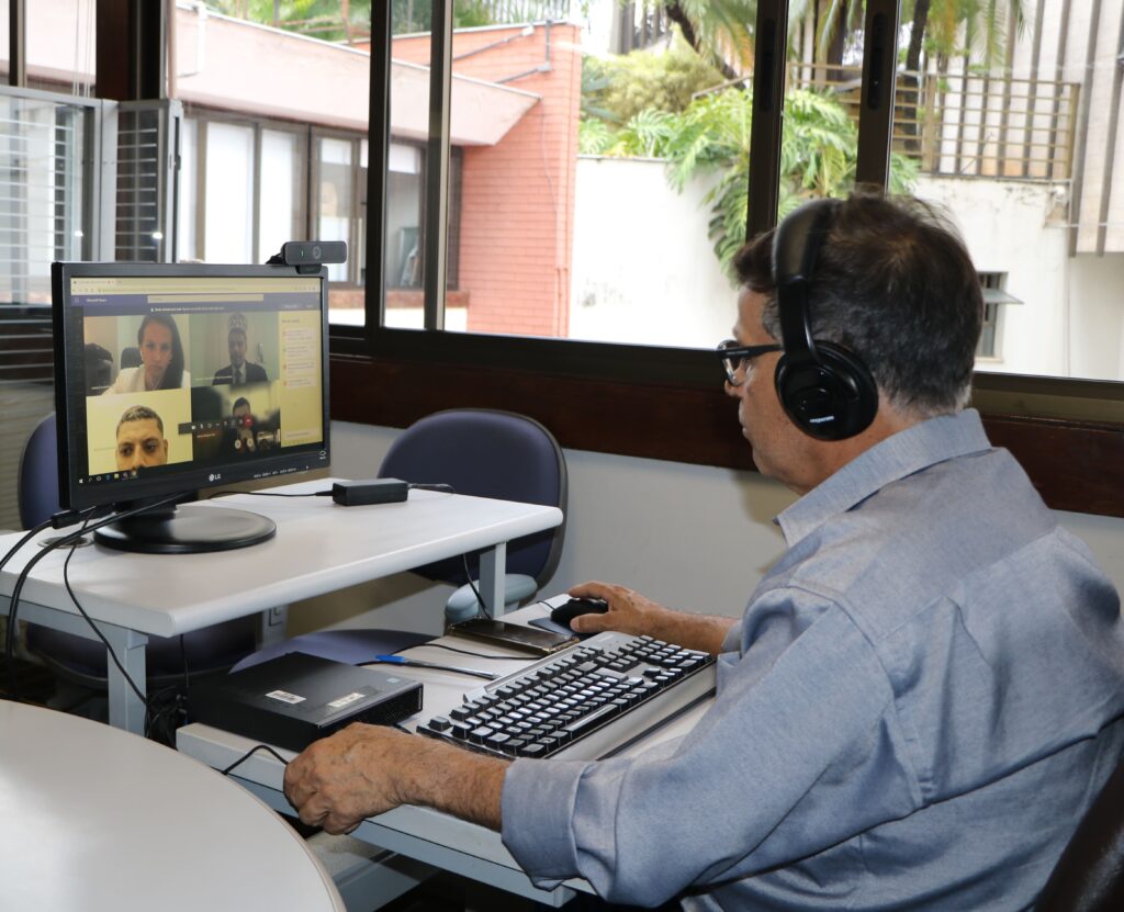 Fotografia colorida homem com fone de ouvido sentado em frente à mesa de computador com a mão no mouse participando de reunião virtual.