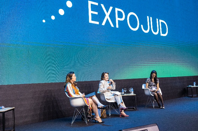 Fotografia com formato arredondado colorida com três mulheres sentadas em palco de evento, uma delas falando ao microfone. 