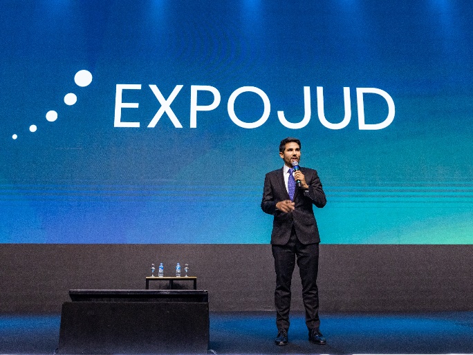 Fotografia horizontal colorida homem em pé no palco do evento falando ao microfone, com nome do evento ao fundo.