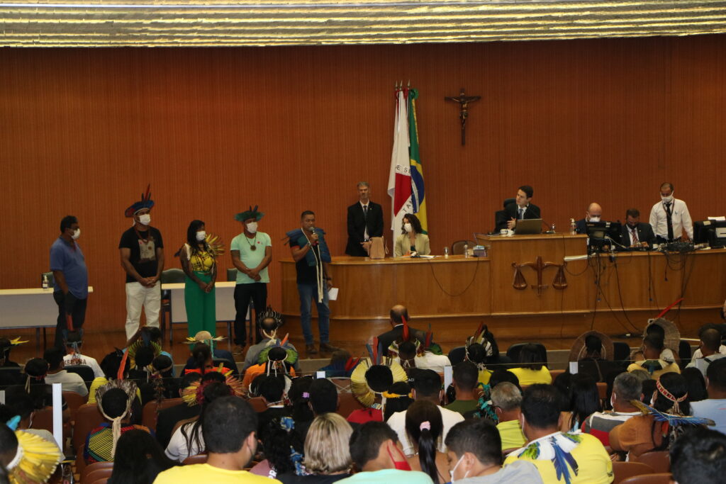Fotografia retangular e colorida em que um indígena fala ao microfone, diante de uma plateia de outros indígenas, numa audiência judicial com o magistrado à direita.