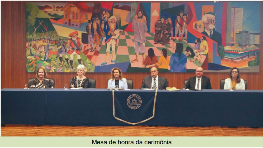 Fotografia retangular e colorida em que seis pessoas se encontram sentadas numa mesa de honra, com uma pintura histórica ao fundo. São quatro mulheres e dois homens, ambos não-negros.