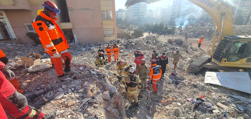 Foto horizontal, colorida, de bombeiros durante o trabalho de resgate, em meio a escombros de prédios.  