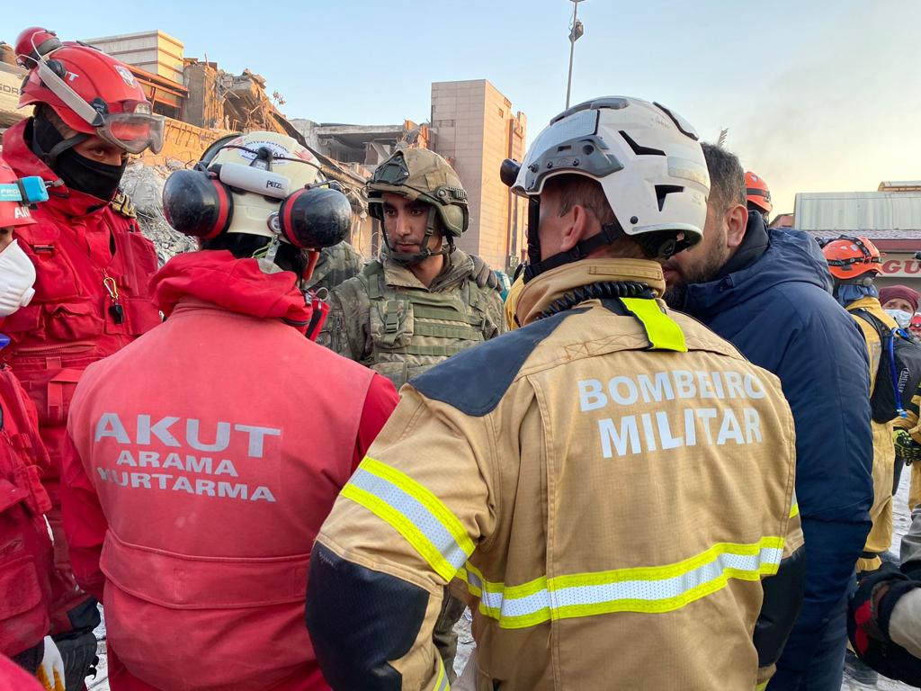 Foto horizontal, colorida, de um grupo de bombeiros conversando. Todos usam trajes e equipamentos de equipes de resgate