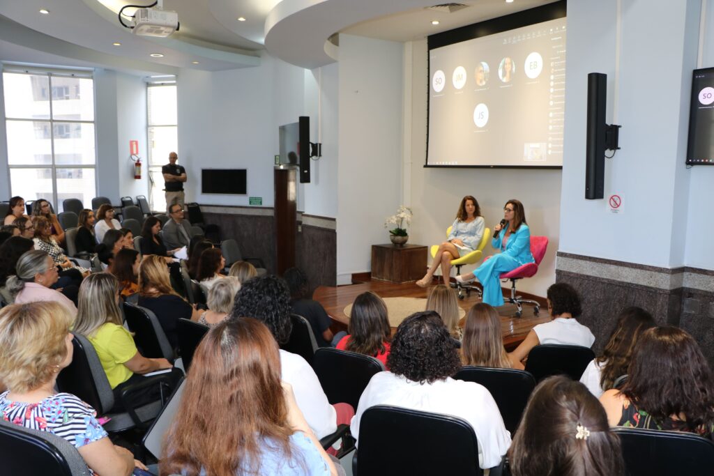 Fotografia retangular e colorida em que duas mulheres não-negras estão sentadas diante de uma plateia majoritariamente feminina, num auditório. Uma das mulheres fala ao microfone.