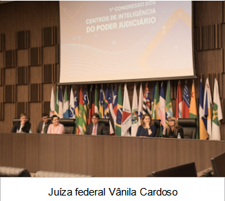 Foto quadrada, colorida, de pessoas sentadas à mesa de honra do evento, enquanto uma mulher de pele branca fala ao microfone.

Legenda: Juíza federal Vânila Cardoso 
