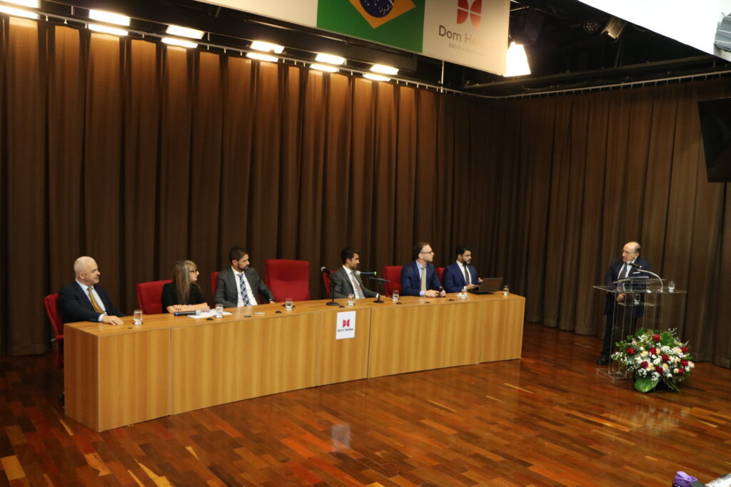 Foto retangular colorida mostra uma mesa de autoridades usando trajes formais. Elas estão sentadas e olham para um homem que profere uma palestra para um auditório.