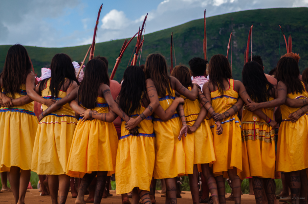 Fotografia retangular e colorida em que  nove mulheres indígenas estão de costas, de braços dados e vestidas.