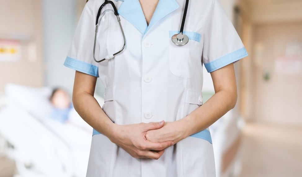 Fotografia retangular colorida de uma enfermeira com roupas brancas e estetoscópio ao redor do pescoço.