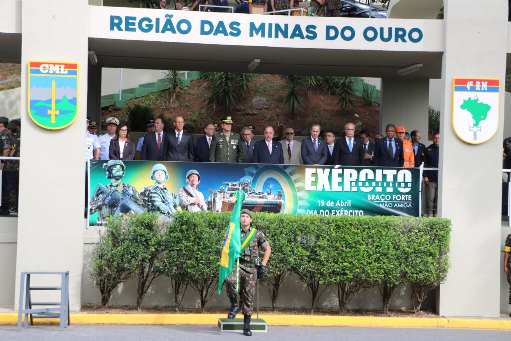 Fotografia colorida vertical com diversas pessoas em uma tribuna e, à frente, um militar segura a bandeira do Brasil. Legenda: Presidente do TRF6 participa da tribuna de honra da solenidade do Dia do Exército.