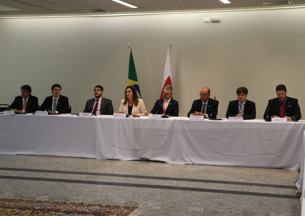 Fotografia retangular e colorida com sete homens e uma mulher ao centro, todos sentados numa mesa, com duas bandeiras (Brasil e Minas Gerais).