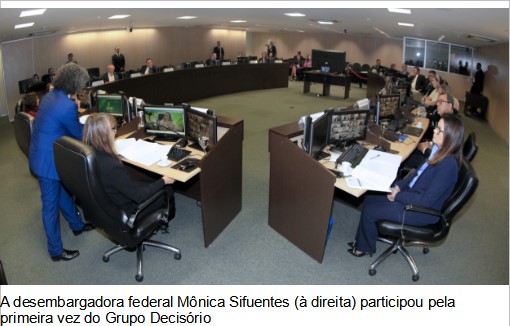Fotografia horizontal colorida de um auditório com uma bancada em forma de círculo onde estão sentados  participantes da reunião. No primeiro plano, aparece a Presidente do TRF6 sentada de lado. No seu lado oposto, aparece a juíza Vânila Cardoso sentada de costas.