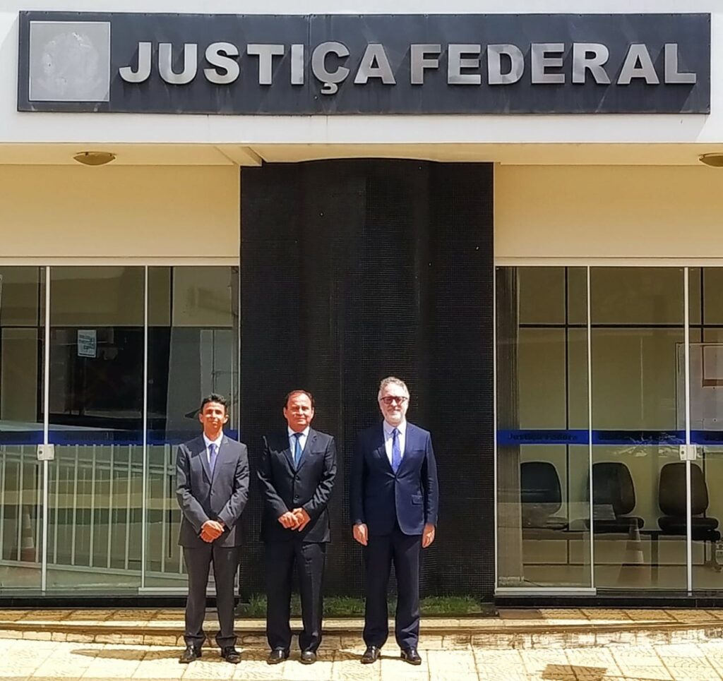 Fotografia quadrada e colorida em que aparecem três homens de terno em frente a um prédio. Acima deles, a inscrição "Justiça Federal".