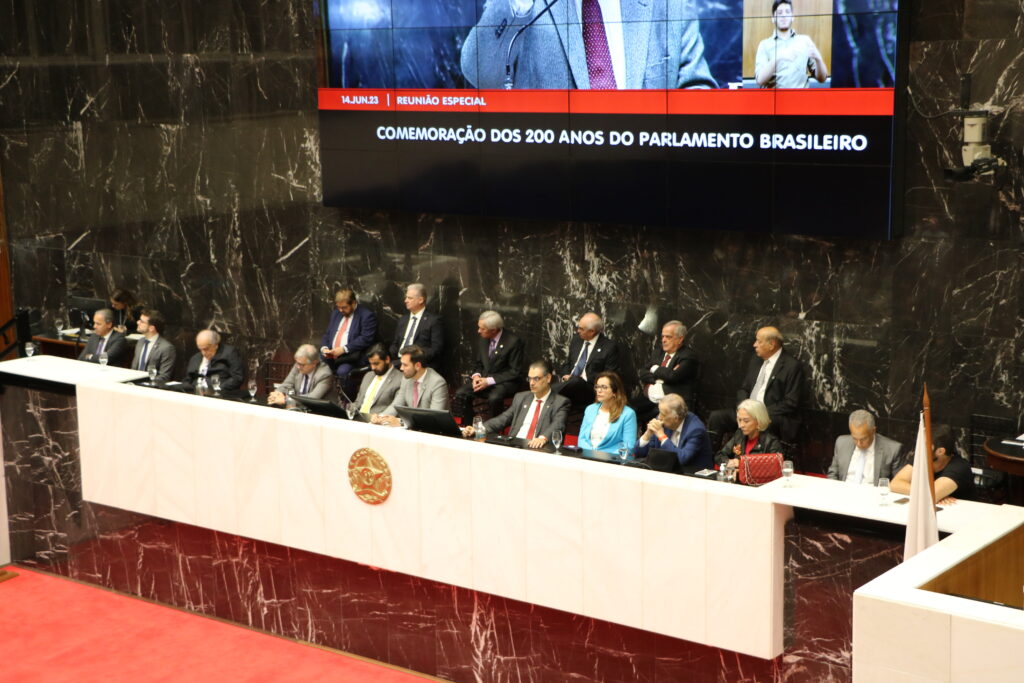 Foto colorida horizontal mostra a mesa do plenário da Assembleia Legislativa com autoridades sentadas em duas fileiras. 