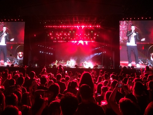 Fotografia colorida e retangular em que aparece uma multidão na frente de um palco com dois telões de cada lado.
