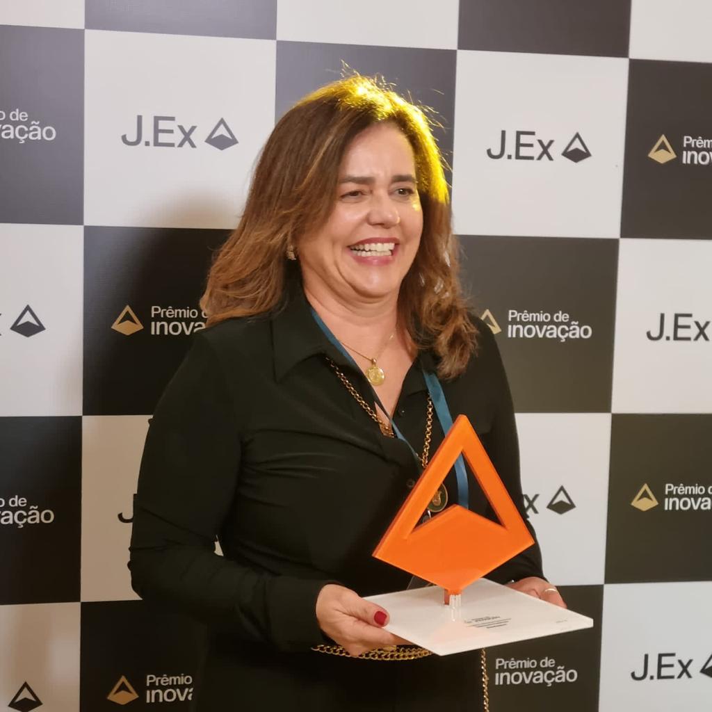 Fotografia colorida vertical em que aparece uma mulher segurando o "Prêmio de Inovação J.Ex", de cor laranja.
