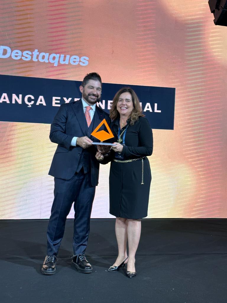 Fotografia colorida vertical em que aparecem um homem e uma mulher segurando o "Prêmio de Inovação J.Ex", de cor laranja.