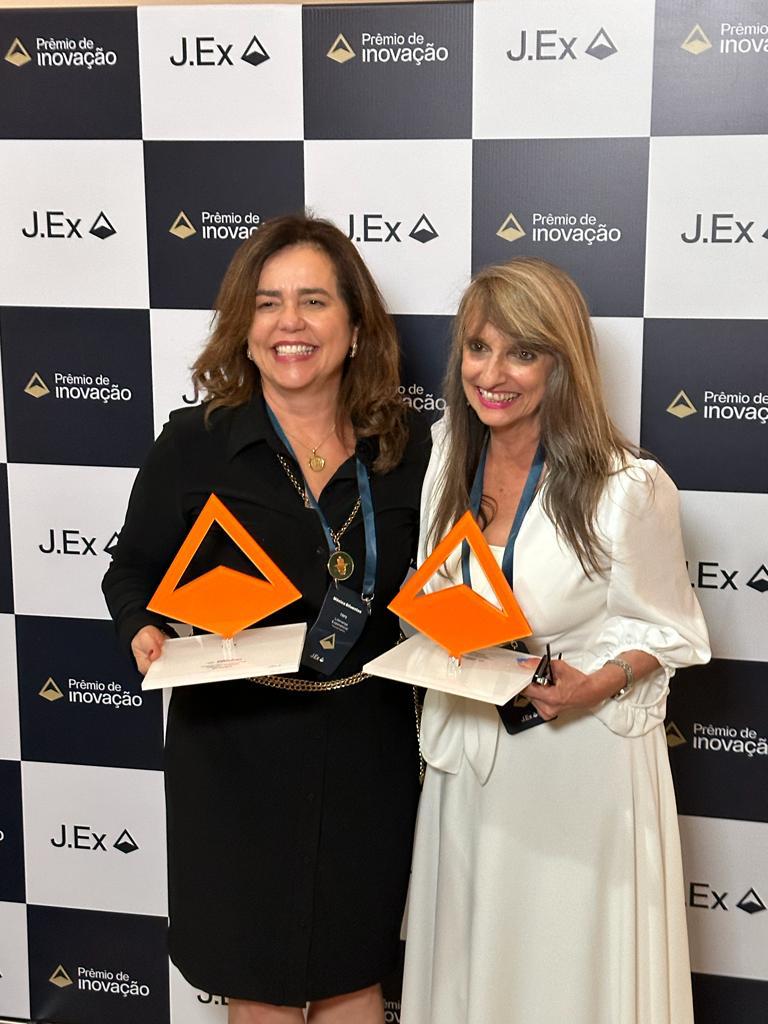 Fotografia colorida vertical em que aparecem duas mulheres segurando o "Prêmio de Inovação J.Ex", de cor laranja.