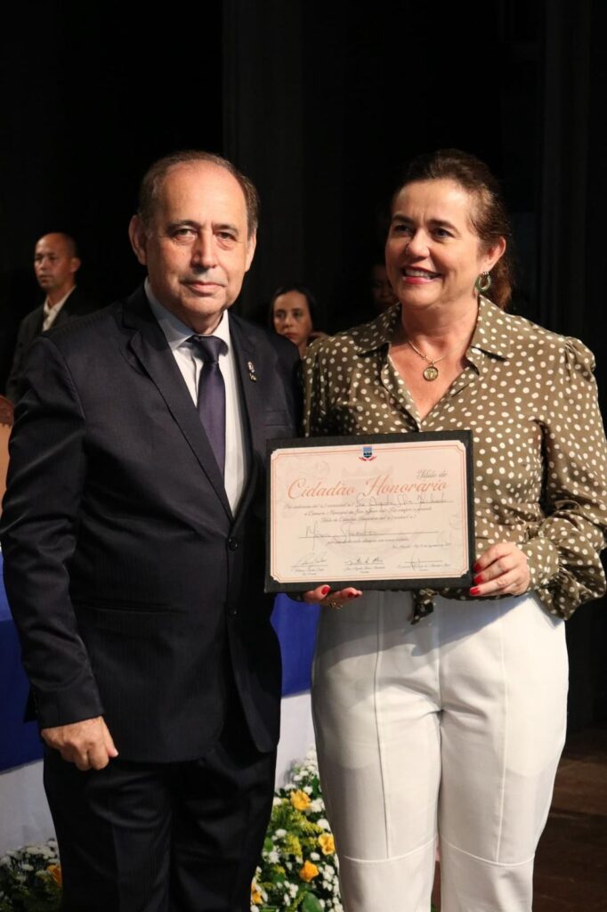 Imagem colorida de uma mulher recebendo uma placa de “Cidadã Honorária” de um homem.