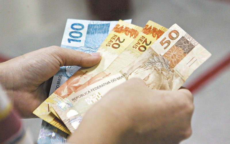 Fotografia colorida das mãos de uma pessoa contando notas de R$50, R$20 e R$100 reais.