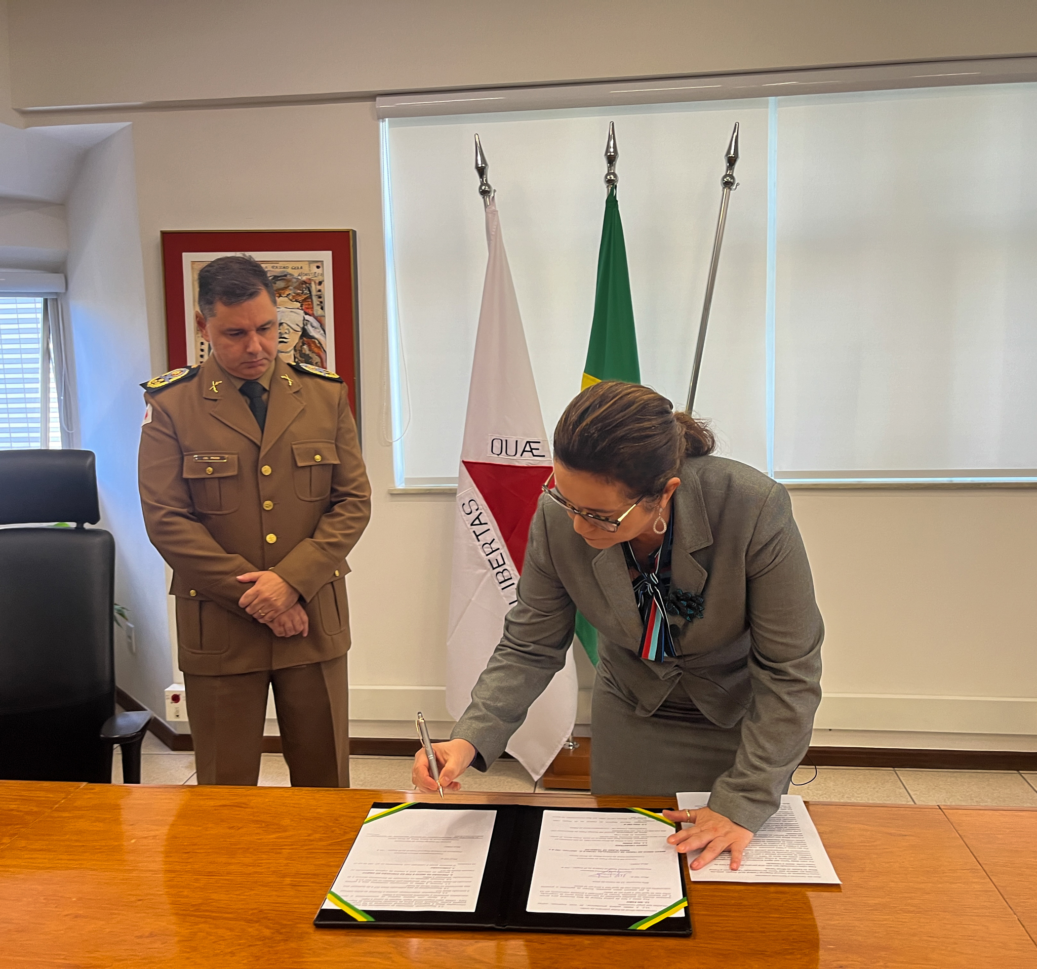 Fotografia colorida de uma mulher de óculos assinando um documento. À direita, o comandante-geral da PMMG observa a assinatura.