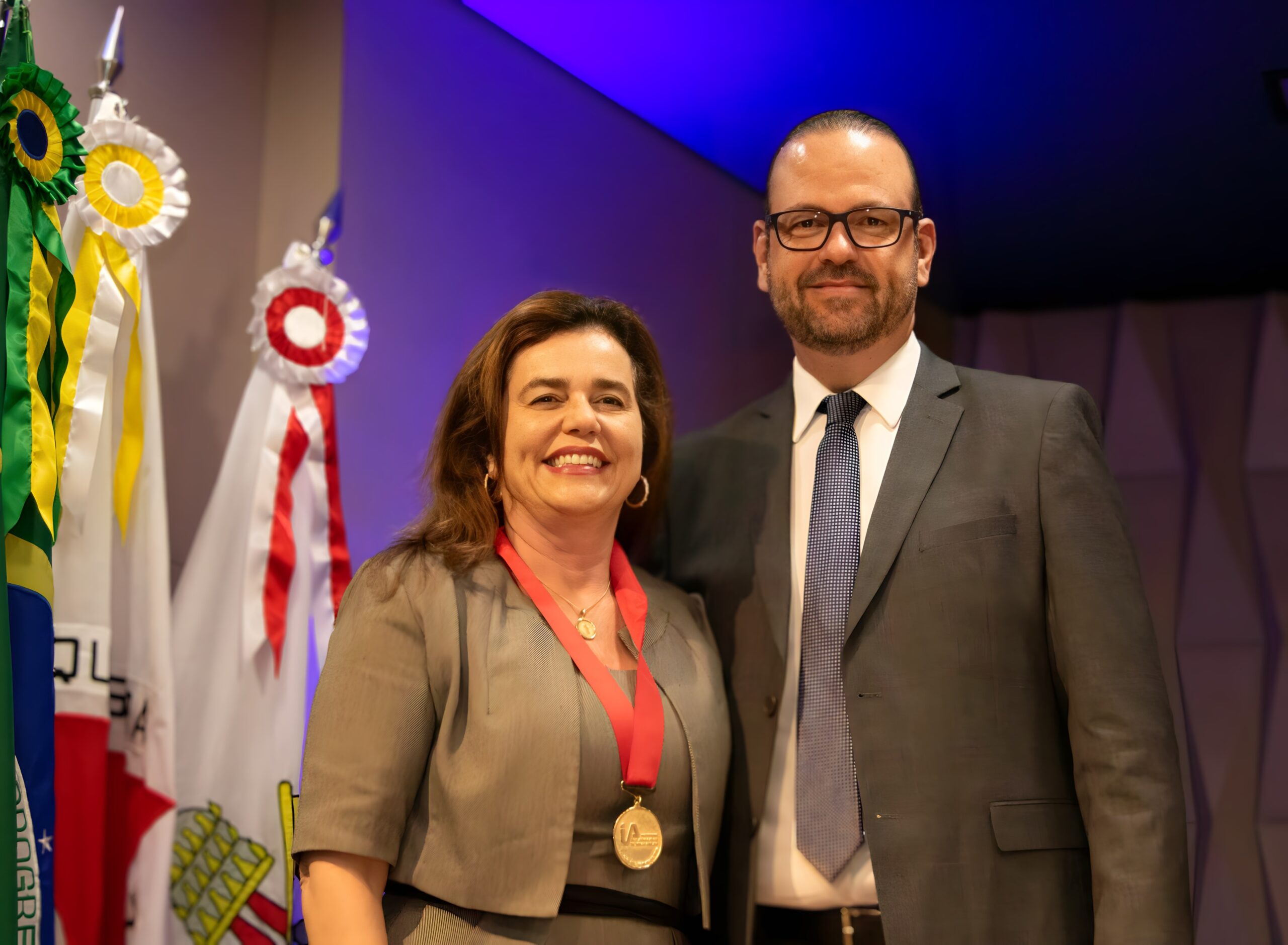 Fotografia colorida de um homem de terno, gravata e óculos e uma mulher com uma medalha do Instituto dos Advogados de Minas Gerais ao redor do pescoço.