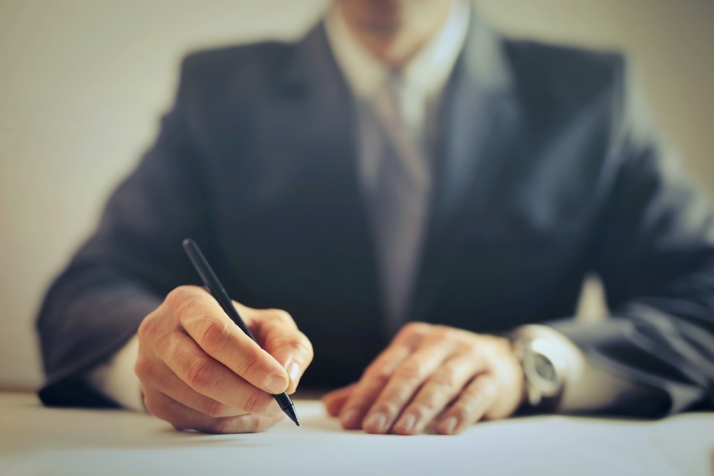 Imagem colorida da mão direita de uma pessoas segurando uma caneta e assinando um documento. 