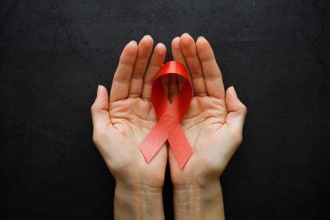 Fotografia colorida e retangular em que duas mãos seguram o laço-símbolo da luta contra a AIDS.