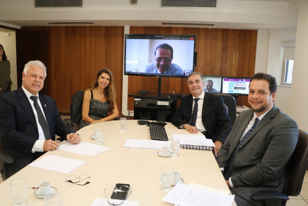Fotografia colorida de uma mesa de reunião com três homens e uma mulher sentados e um homem participando virtualmente, através da tela de um monitor.
