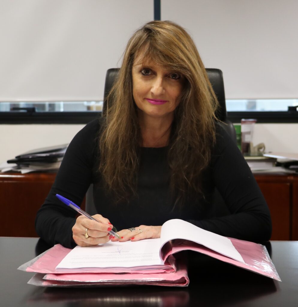 Fotografia colorida de uma mulher com uma caneta nas mãos assinando alguns documentos.
