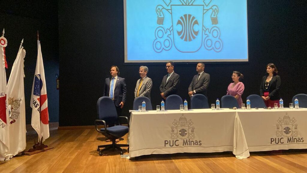 Fotografia colorida de 4 homens e duas mulheres de pé, atrás de uma mesa de evento, voltados para as bandeiras que estão posicionadas ao lado da mesa.