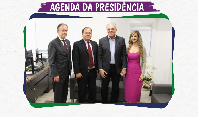 A imagem mostra quatro pessoas em pé, posando para uma foto em um ambiente de escritório. Três homens de terno e gravata, e uma mulher com vestido rosa e um casaco claro. Ao fundo, há móveis de escritório, incluindo mesas com computadores e cadeiras, e uma decoração com planta ao lado.