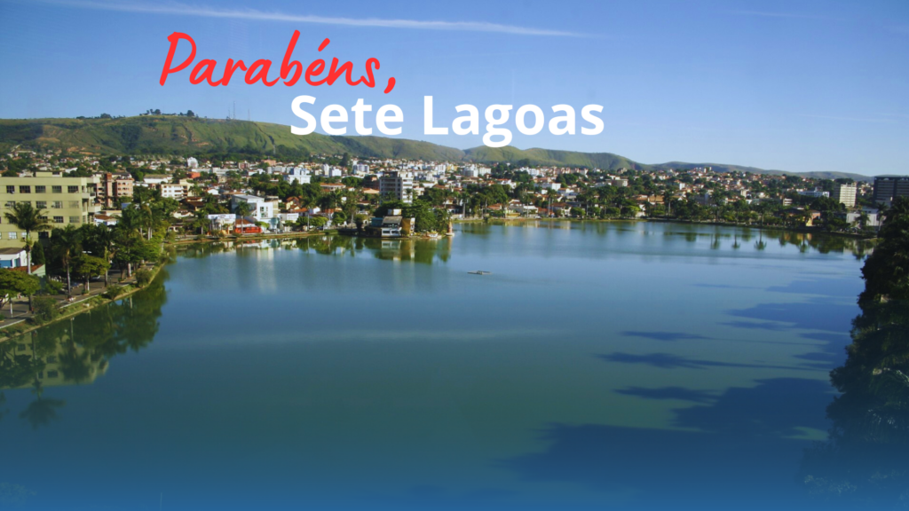 A imagem apresenta uma paisagem urbana da cidade de Sete Lagoas, destacando uma bela lagoa no centro, com água calma refletindo o céu azul. Ao fundo, há construções urbanas e colinas verdes, compondo um cenário harmonioso.

O texto está sobreposto na parte inferior da imagem com as palavras:

"Parabéns," em vermelho, com uma fonte cursiva.
"Sete Lagoas" em branco, com uma fonte mais simples e em destaque.