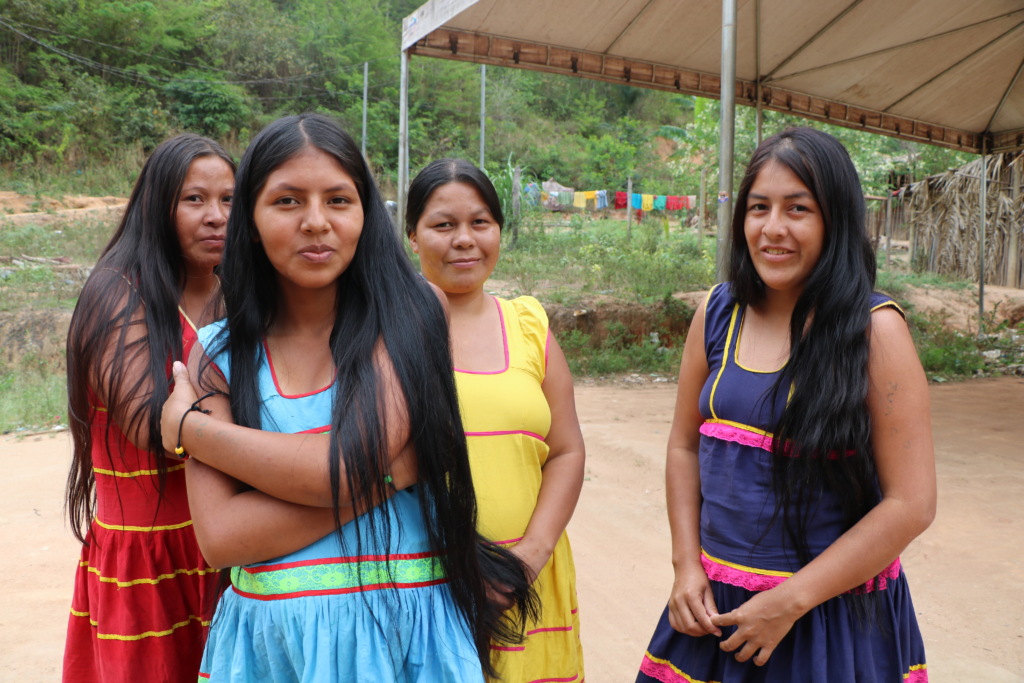 Fotografia colorida de um grupo de pessoas reunidas em um ambiente comunitário, para uma reunião. Dois homens estão em destaque à frente, um ao lado do outro, um deles está falando. Há várias pessoas ao redor deles, incluindo mulheres e crianças sentadas e em pé, com um cachorro deitado no banco ao fundo. 