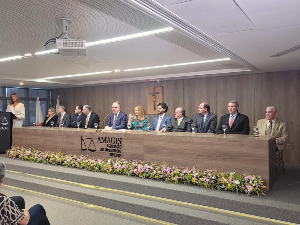 A imagem retrata uma cerimônia formal com várias pessoas em pé atrás de uma longa mesa de madeira. A mesa está decorada com um arranjo de flores na parte inferior. Na frente da mesa, está o logotipo e o nome da AMAGIS (Associação dos Magistrados Mineiros). As pessoas estão alinhadas, aparentando ser figuras públicas ou autoridades, todas em trajes formais, em um ambiente fechado, iluminado e bem organizado. No teto, há luminárias embutidas, e o espaço apresenta um acabamento moderno.