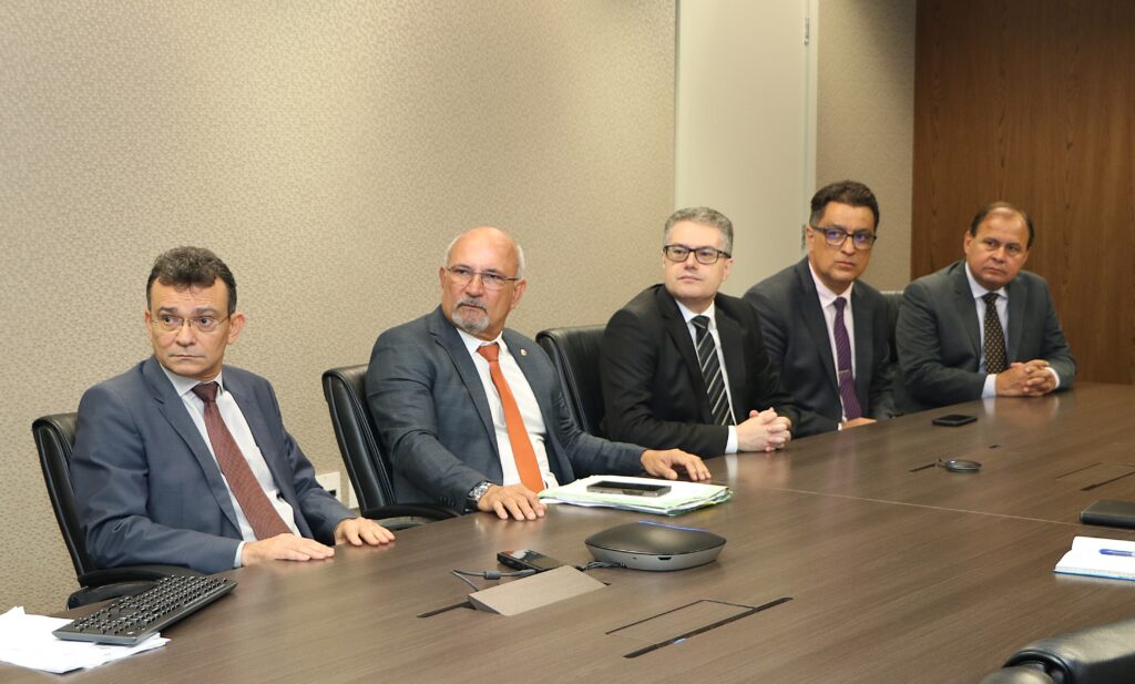 Nesta fotografia há cinco homens de terno sentados atrás de uma mesa. 