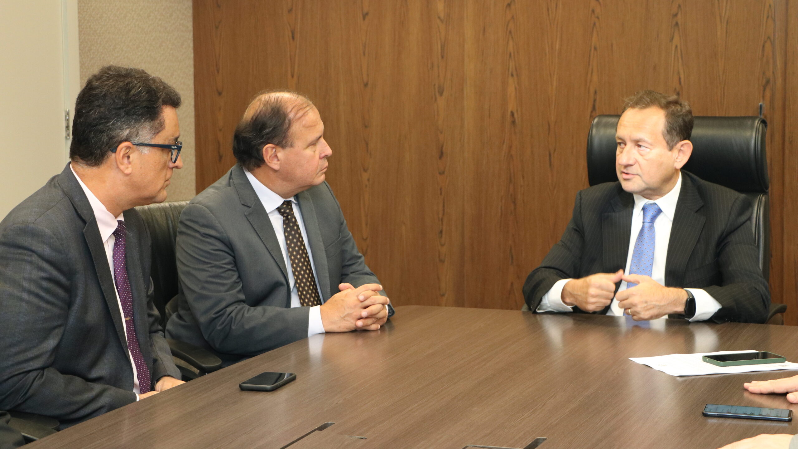 Nesta imagem há três homens brancos de terno e gravata sentados atrás de uma mesa. Eles estão conversando entre si. 