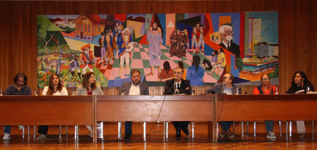 A imagem exibe um grupo de oito pessoas, incluindo homens e mulheres, sentadas atrás de uma longa mesa de madeira em um palco ou plataforma elevada. Ao fundo, um grande mural colorido com figuras abstratas e cenas urbanas serve como pano de fundo.

O piso do palco é de madeira e a iluminação é artificial.