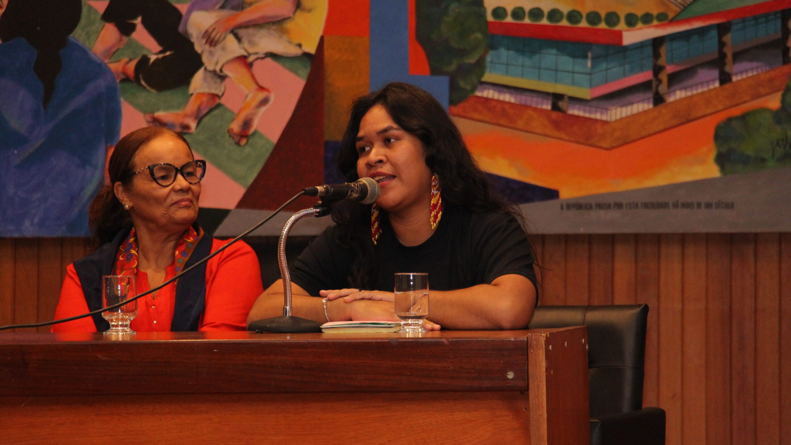 A imagem apresenta duas mulheres sentadas a uma mesa de madeira escura, com um grande mural colorido ao fundo.

À esquerda, uma mulher de óculos e cabelo preso, com um casaco vermelho sobre uma blusa escura, está sentada, com expressão séria. À direita, uma mulher mais jovem, com longos cabelos escuros e brincos, está falando em um microfone que está à sua frente. Suas mãos estão sobre a mesa e há um copo de água perto dela.