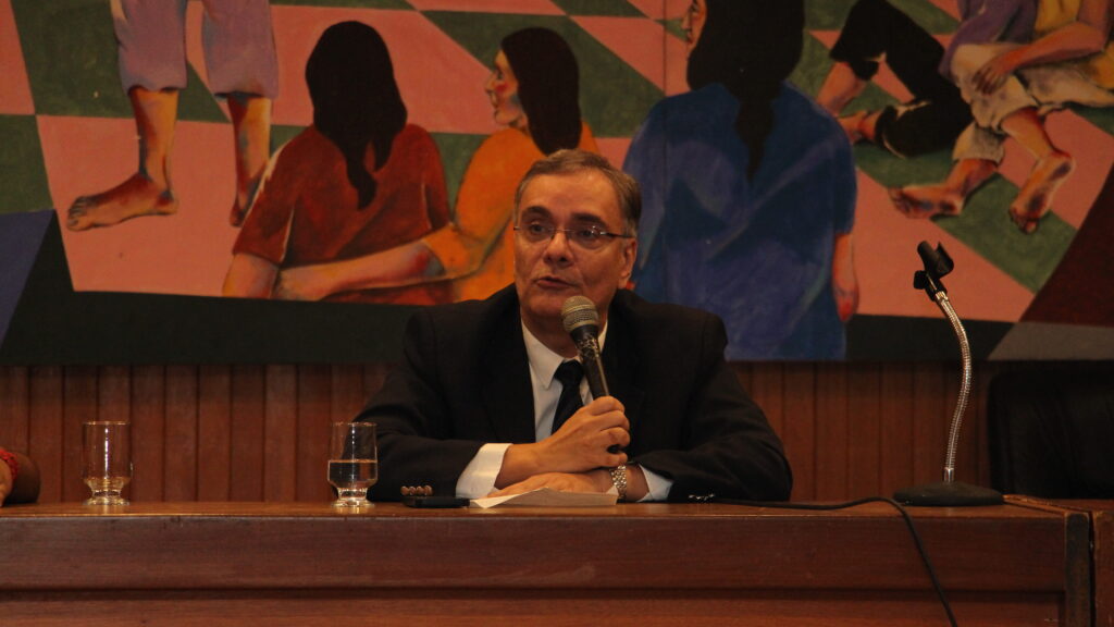 A imagem centraliza um homem de meia-idade, com cabelos grisalhos e óculos, sentado a uma mesa de madeira escura. Ele está segurando um microfone e com a boca ligeiramente aberta. Há dois copos de água sobre a mesa, um à esquerda e outro à direita do microfone. Ao fundo, há um grande mural artístico com figuras abstratas e coloridas. O ambiente é formal.