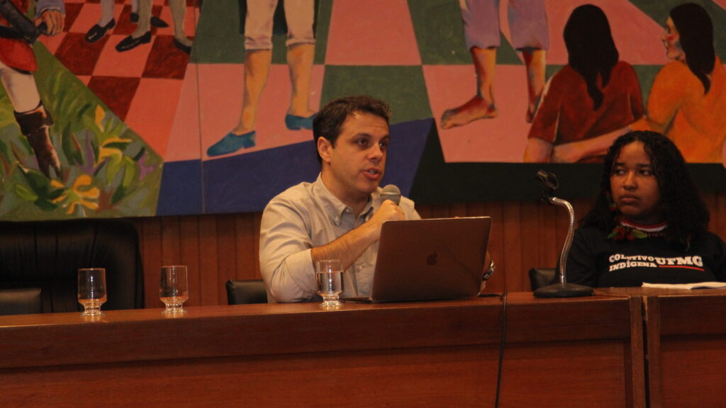 A imagem exibe duas pessoas sentadas a uma mesa de madeira escura, com um grande mural colorido ao fundo.

À esquerda, um homem está sentado com um laptop aberto à sua frente e um copo de água ao lado. Ele segura um microfone e parece estar falando. À direita dele, uma mulher com longos cabelos escuros está sentada, olhando para o homem, com um microfone à sua frente e um copo de água ao lado.