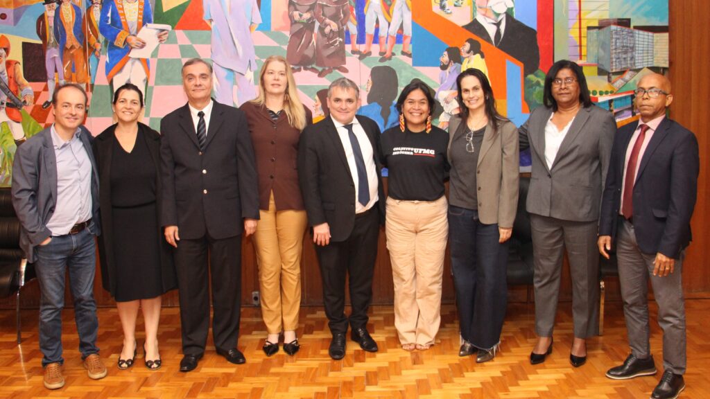 A imagem mostra um grupo de nove pessoas, incluindo homens e mulheres, posando em um ambiente interno com um grande mural artístico ao fundo. O mural apresenta figuras humanas e formas geométricas em cores vibrantes como azul, roxo, verde, amarelo e vermelho.

Da esquerda para a direita, os indivíduos são:

Um homem de cabelos escuros vestindo um blazer cinza sobre uma camisa clara e calças jeans. Ele sorri.
Uma mulher com cabelos escuros presos, vestindo um vestido preto sem mangas. Ela sorri.
Um homem de cabelos grisalhos, usando um terno escuro com camisa branca e gravata preta.
Uma mulher loira com cabelos compridos, vestindo um casaco marrom e calças beges.
Um homem de meia-idade com cabelos grisalhos, vestindo um terno preto com camisa branca e gravata azul.
Uma mulher com cabelos escuros, vestindo uma camiseta preta com uma estampa branca e calças beges. Ela sorri.
Uma mulher de cabelos longos e escuros, usando óculos e um blazer cáqui sobre uma blusa escura e jeans. Ela sorri.
Uma mulher de cabelos curtos e escuros, usando óculos e um terno cinza escuro sobre uma blusa branca.
Um homem com óculos, vestindo um terno azul-marinho e gravata vermelha. Ele sorri.
