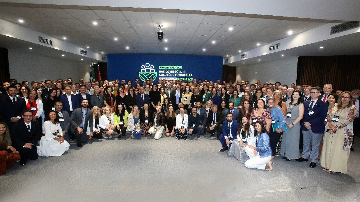 A imagem exibe um grande grupo de pessoas reunidas em um auditório ou salão de eventos, posando para uma foto.

A maioria das pessoas está de pé, e algumas na frente estão agachadas ou sentadas, formando um semicírculo. No geral, os participantes estão vestidos com trajes sociais ou esporte fino.

Ao fundo, há um painel azul grande com a inscrição "ENCONTRO ANUAL DAS COMISSÕES DE SOLUÇÕES FUNDIÁRIAS" e um logotipo que inclui figuras humanas estilizadas. A iluminação do local é bem distribuída.