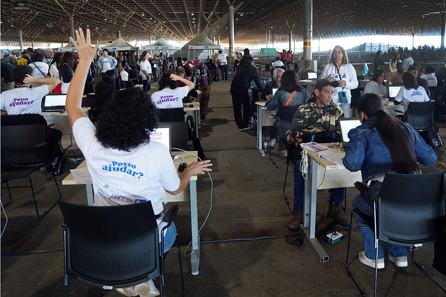 A imagem retrata um grande evento em um espaço amplo, com muitas pessoas e mesas de atendimento. O evento parece ser um mutirão de serviços ou assistência.

Em primeiro plano, há várias mesas com computadores. Uma pessoa com cabelos cacheados e uma camiseta branca com a frase "Posso ajudar?" nas costas está sentada em frente a um computador, com um dos braços levantado, como se estivesse chamando alguém ou pedindo atenção. Outra pessoa, sentada na mesa à direita, está usando um computador e parece estar auxiliando um usuário.

Ao fundo, há inúmeras outras mesas de atendimento com pessoas sentadas, algumas usando computadores. Tendas ou estruturas temporárias podem ser vistas ao longo do espaço. O ambiente tem um teto alto com estrutura metálica aparente.

Abaixo da imagem, há uma legenda: "Mutirão PopRuaJud - Foto: G. Dettmar/Ag. CNJ".