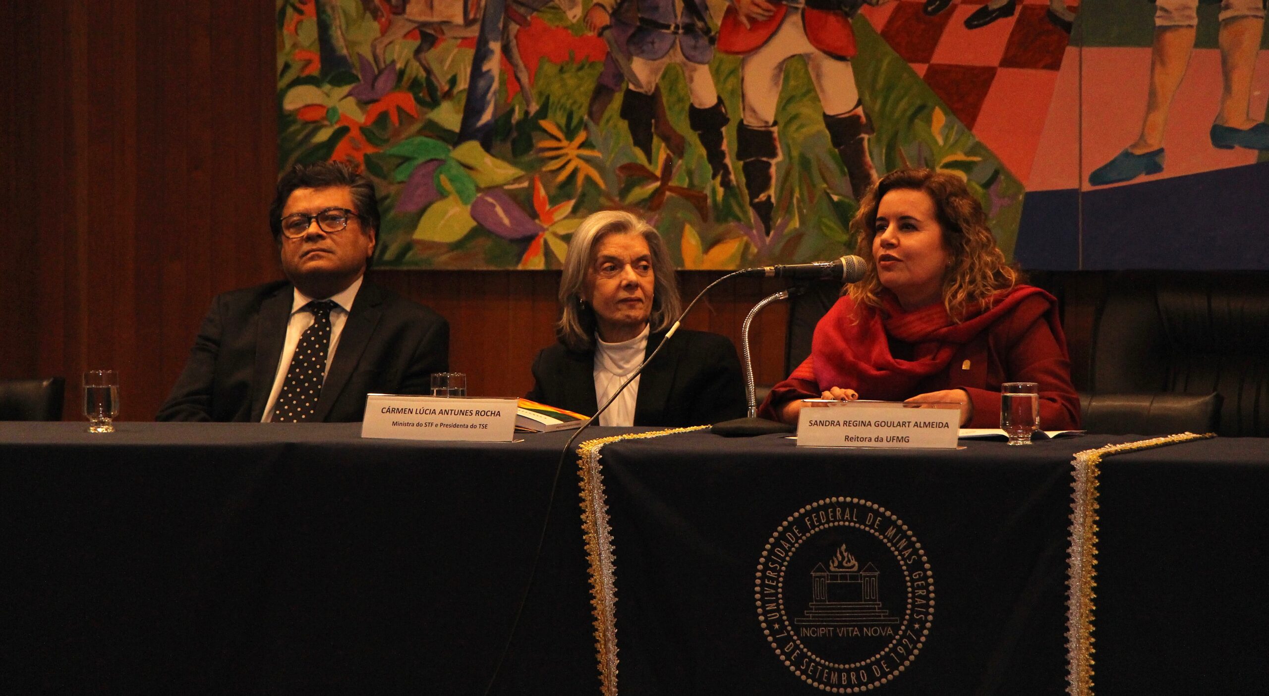 A imagem mostra três pessoas sentadas em uma mesa coberta com um tecido azul escuro.

A pessoa à esquerda é um homem de óculos, vestindo um terno escuro com uma camisa branca e uma gravata listrada. A pessoa ao centro é uma mulher com cabelos grisalhos, vestindo uma blusa escura com gola alta branca. A pessoa à direita é uma mulher com cabelos cacheados castanhos claros, usando um agasalho vermelho.

Na mesa, há algumas placas de identificação com nomes e títulos. Um copo com água pode ser visto em frente ao homem. A frente da mesa é decorada com uma fita dourada e há um emblema circular no centro.