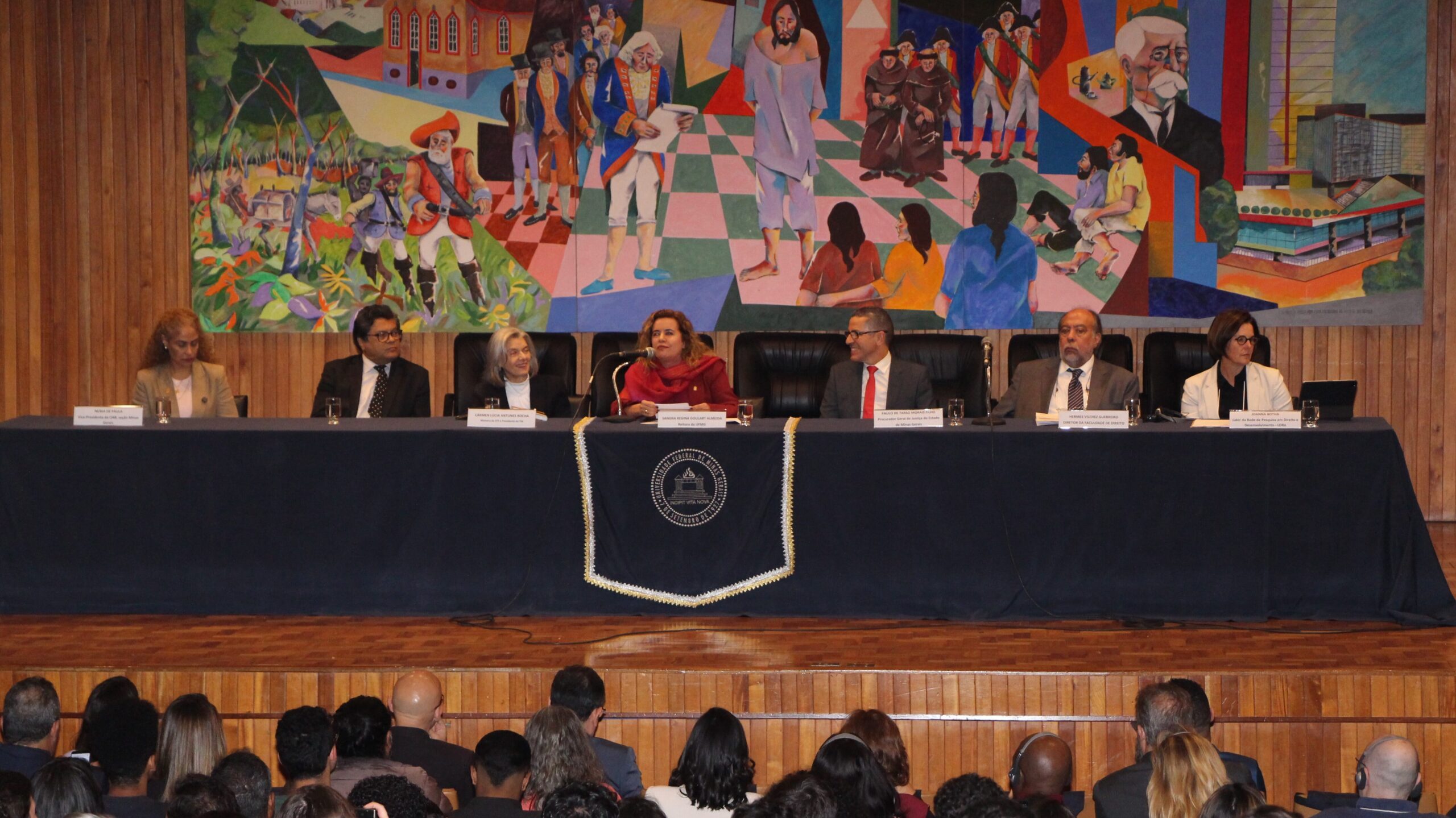 A imagem mostra um grupo de sete pessoas, três homens e quatro mulheres, em um ambiente interno formal. Eles estão assentados, lado a lado. Ao fundo, um quadro gigante é visível.

Em primeiro plano, vê-se uma plateia numerosa, com muitas pessoas sentadas em cadeiras, olhando para a frente.