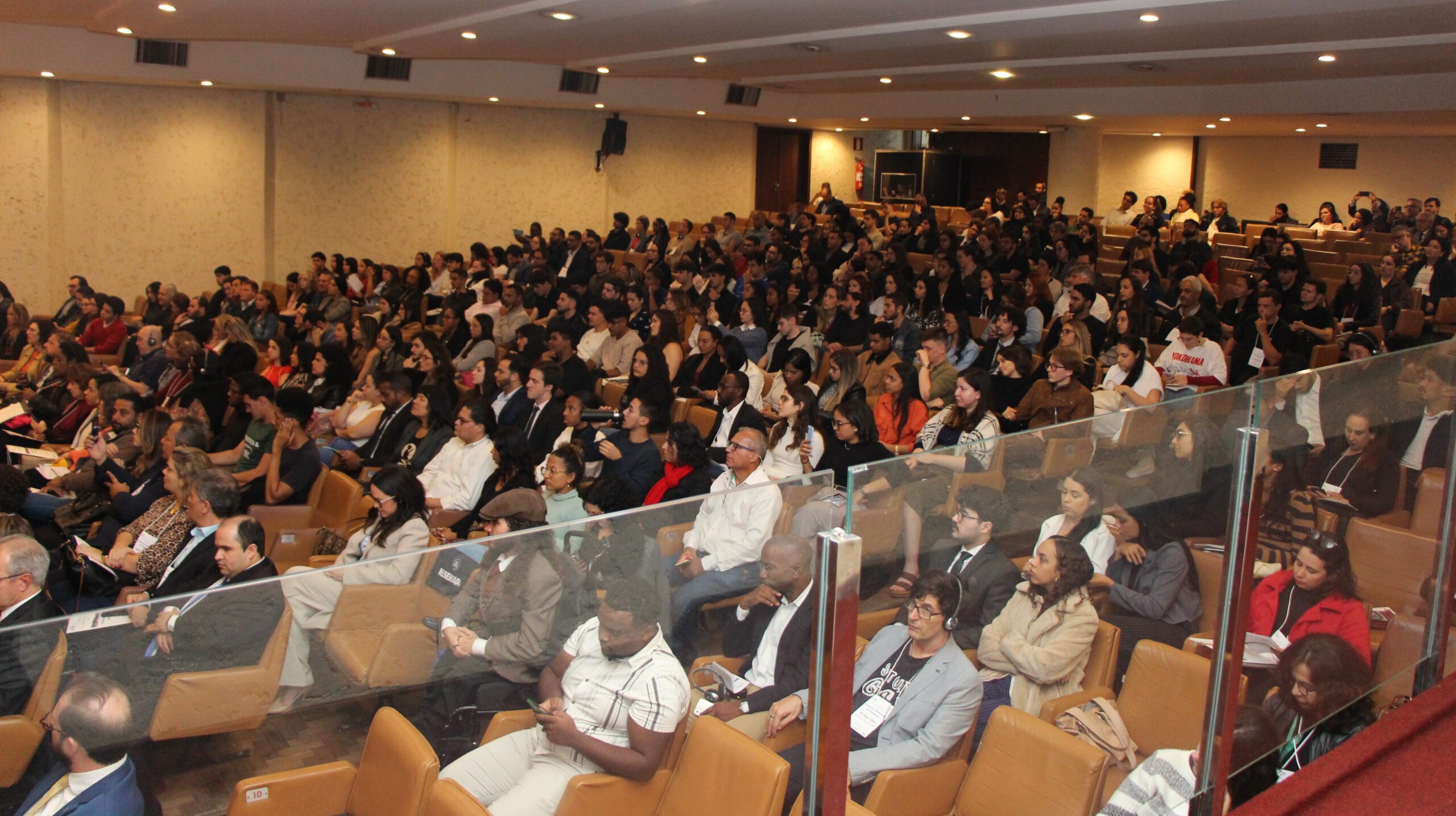 A imagem mostra uma grande plateia de pessoas sentadas em um auditório. As cadeiras são de tonalidade marrom-claro ou bege. Há muitas pessoas de diversas idades, a maioria delas está olhando para frente.

Há uma barreira de vidro transparente que separa as primeiras filas da visão do observador. No fundo do auditório, é possível ver mais fileiras de cadeiras e algumas pessoas.