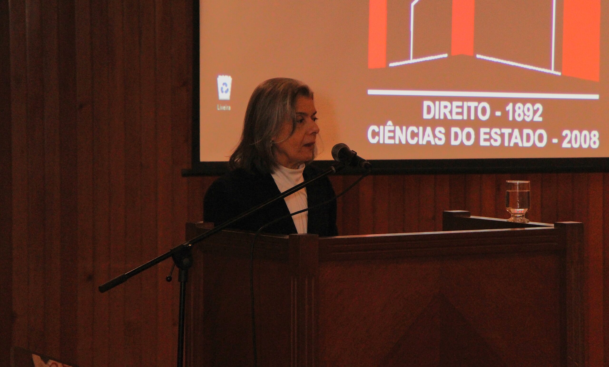 A imagem mostra uma mulher de cabelos grisalhos em pé atrás de um púlpito de madeira. Ela está falando em um microfone que está montado no púlpito.

A mulher veste uma blusa escura com uma gola alta branca por baixo.

Ao fundo, há uma parede de madeira escura e, acima do púlpito, uma projeção em tela exibe texto em letras maiúsculas: "DIREITO - 1892" e "CIÊNCIAS DO ESTADO - 2008". A projeção também contém elementos gráficos em tons de laranja e branco. À direita da mulher, sobre o púlpito, há um copo com água.